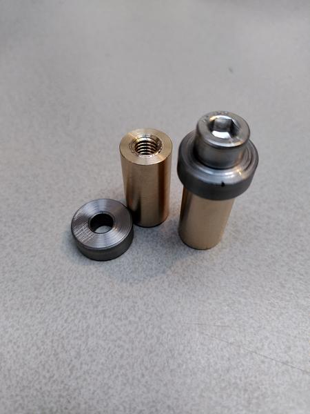 Two brass cylinders stood on end on a mottle grey table. The one on the
right has a short but wide piece of steel on top and fixed in place with a
hex bolt. The other has the steel piece next to it on the table.